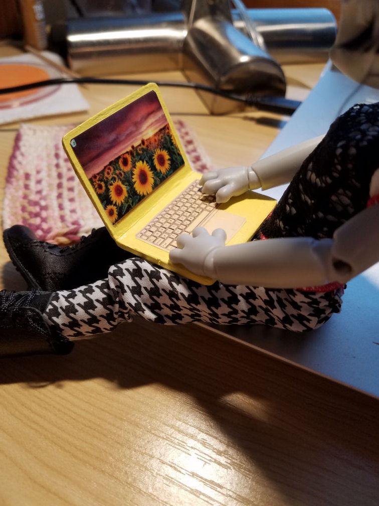 Looking over the shoulder of a grey Hujoo Phoebe Mouse doll sitting on the edge of a silver laptop with a doll sized pale yellow laptop open on her lap. The laptop has a dramatic photo of a field of sunflowers under a stormy sunset for a wallpaper, a recycle bin, and a taskbar, but no open applications.